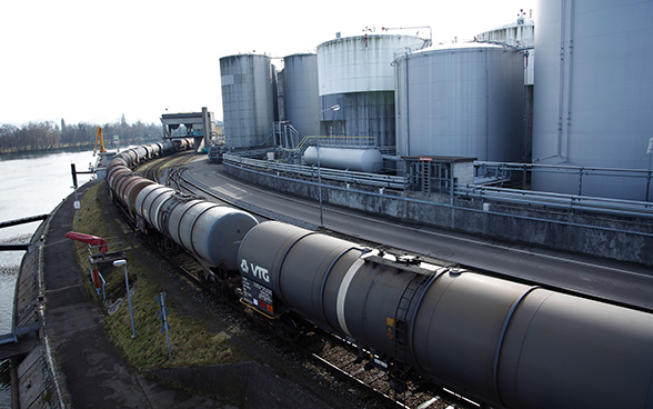 Des wagons-citernes gris-marron empruntent une voie entre le Rhin et des silos.